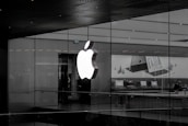 A large illuminated apple logo on the glass facade of a store. Inside, various Apple products such as laptops are displayed on tables with illuminated screens. The store interior appears modern and minimalistic.