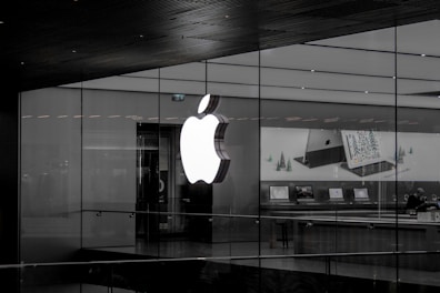 A large illuminated apple logo on the glass facade of a store. Inside, various Apple products such as laptops are displayed on tables with illuminated screens. The store interior appears modern and minimalistic.