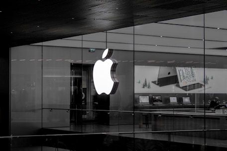 A large illuminated apple logo on the glass facade of a store. Inside, various Apple products such as laptops are displayed on tables with illuminated screens. The store interior appears modern and minimalistic.