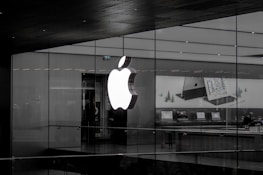 A large illuminated apple logo on the glass facade of a store. Inside, various Apple products such as laptops are displayed on tables with illuminated screens. The store interior appears modern and minimalistic.