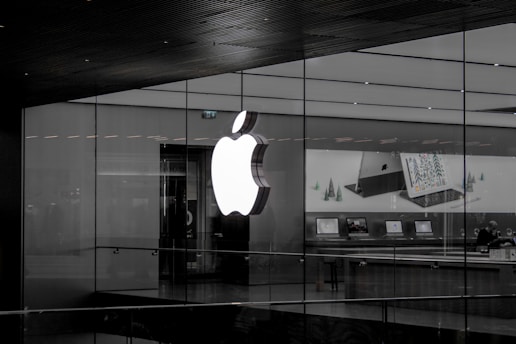 A large illuminated apple logo on the glass facade of a store. Inside, various Apple products such as laptops are displayed on tables with illuminated screens. The store interior appears modern and minimalistic.