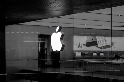 A large illuminated apple logo on the glass facade of a store. Inside, various Apple products such as laptops are displayed on tables with illuminated screens. The store interior appears modern and minimalistic.