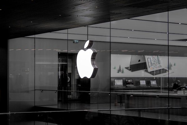A large illuminated apple logo on the glass facade of a store. Inside, various Apple products such as laptops are displayed on tables with illuminated screens. The store interior appears modern and minimalistic.