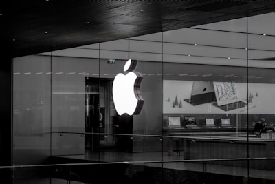 A large illuminated apple logo on the glass facade of a store. Inside, various Apple products such as laptops are displayed on tables with illuminated screens. The store interior appears modern and minimalistic.