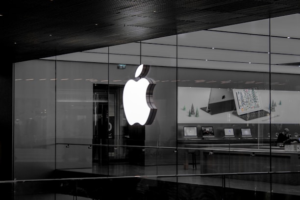 A large illuminated apple logo on the glass facade of a store. Inside, various Apple products such as laptops are displayed on tables with illuminated screens. The store interior appears modern and minimalistic.