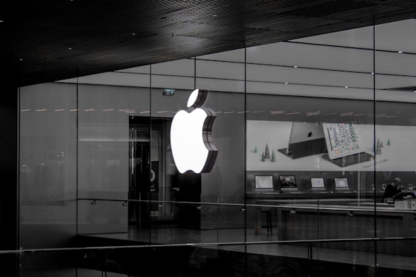 A large illuminated apple logo on the glass facade of a store. Inside, various Apple products such as laptops are displayed on tables with illuminated screens. The store interior appears modern and minimalistic.