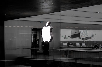A large illuminated apple logo on the glass facade of a store. Inside, various Apple products such as laptops are displayed on tables with illuminated screens. The store interior appears modern and minimalistic.