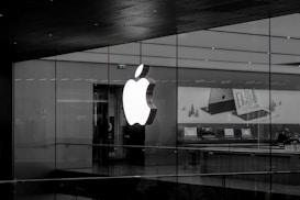 A large illuminated apple logo on the glass facade of a store. Inside, various Apple products such as laptops are displayed on tables with illuminated screens. The store interior appears modern and minimalistic.