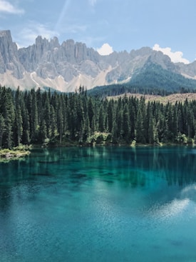 A serene mountain landscape featuring a clear blue lake surrounded by dense green pine trees. In the background, rugged grey mountains rise under a bright blue sky with scattered white clouds.