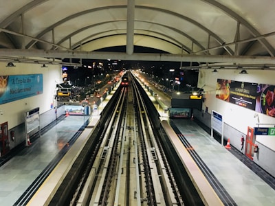 A modern, illuminated metro station with empty tracks stretches into the distance under an arched roof. The station is clean and well-maintained with advertisements on the walls. Bright lights illuminate the tracks as they recede, extending into a night cityscape visible through openings in the background.