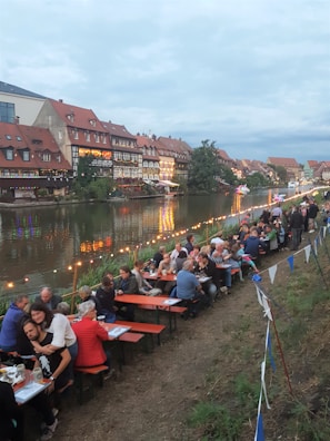 A scenic riverside setting with people dining at picnic-style tables, surrounded by festive string lights and vibrant, historic buildings with red rooftops along the water. The atmosphere appears casual and social, with groups of people engaging in conversation and enjoying themselves. The sky is overcast, creating a calming ambiance.