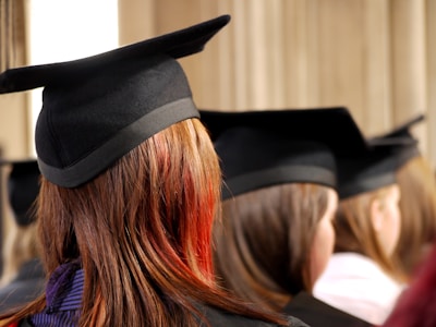 A group of graduates wearing black mortarboards and gowns, seated closely together with their backs turned. The focus is primarily on one individual with long, straight brown hair with a reddish tint, creating a sense of anticipation and celebration.