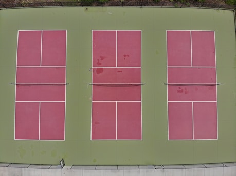 Three rectangular tennis courts with green borders and red playing surfaces viewed from above. The courts are arranged side by side, each divided by a net, with clear white lines marking the boundaries.