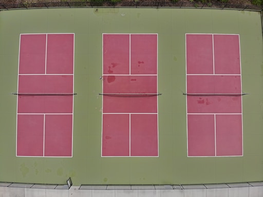 Three rectangular tennis courts with green borders and red playing surfaces viewed from above. The courts are arranged side by side, each divided by a net, with clear white lines marking the boundaries.