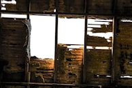 A damaged roof with missing and rotting wooden planks, allowing bright light to shine through. The wooden beams and panels exhibit significant decay and wear, with uneven textures and shadows.