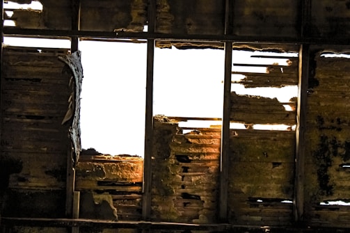A damaged roof with missing and rotting wooden planks, allowing bright light to shine through. The wooden beams and panels exhibit significant decay and wear, with uneven textures and shadows.