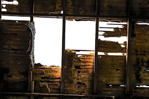 A damaged roof with missing and rotting wooden planks, allowing bright light to shine through. The wooden beams and panels exhibit significant decay and wear, with uneven textures and shadows.