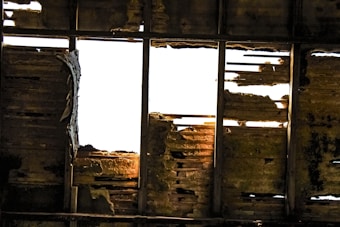 A damaged roof with missing and rotting wooden planks, allowing bright light to shine through. The wooden beams and panels exhibit significant decay and wear, with uneven textures and shadows.