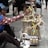 A vendor wearing a patterned shirt and a blue cap is sitting on a sidewalk, arranging traditional handmade toys displayed on a small cart. The toys consist of wooden animal figures and colorful decorations made from bamboo and other natural materials. A few people are seated in the background.