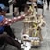 A vendor wearing a patterned shirt and a blue cap is sitting on a sidewalk, arranging traditional handmade toys displayed on a small cart. The toys consist of wooden animal figures and colorful decorations made from bamboo and other natural materials. A few people are seated in the background.
