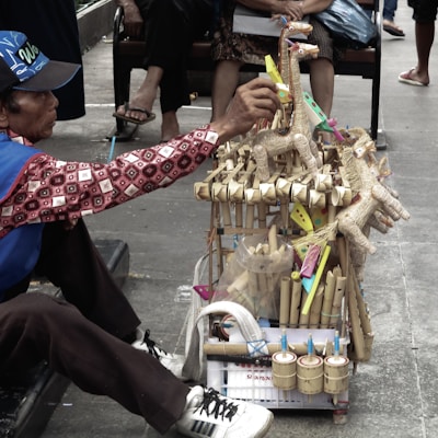 A vendor wearing a patterned shirt and a blue cap is sitting on a sidewalk, arranging traditional handmade toys displayed on a small cart. The toys consist of wooden animal figures and colorful decorations made from bamboo and other natural materials. A few people are seated in the background.