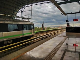 A train station platform with a train stopped on the tracks. The platform is covered with a roof and has a reflective surface, showing the time as 16:14 on a digital clock. The sky is overcast, suggesting the possibility of rain.