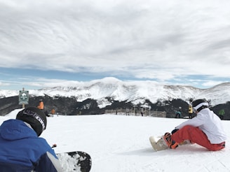 A snowy mountain landscape with several people preparing to snowboard. In the foreground, two individuals are seated on the snow, adjusting their gear. The background features a range of snow-covered peaks under a partly cloudy sky.