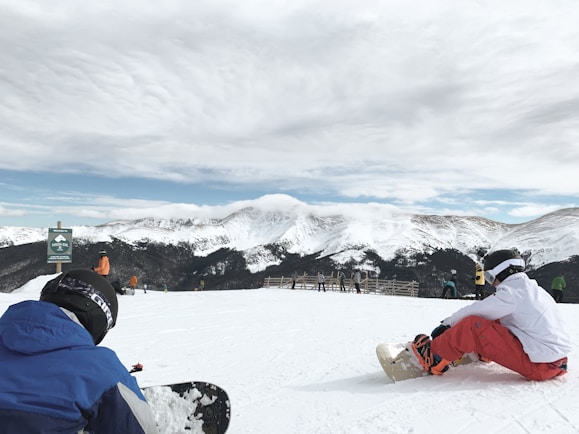 A snowy mountain landscape with several people preparing to snowboard. In the foreground, two individuals are seated on the snow, adjusting their gear. The background features a range of snow-covered peaks under a partly cloudy sky.