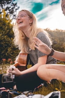 A person with long blonde hair and glasses is sitting on grass and playing a guitar. The background includes lush green trees and a bright sky creating a warm and cheerful atmosphere. Another person is partially visible, adding to a sense of community and joy.
