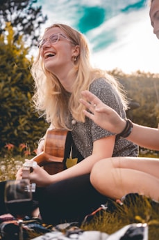 A person with long blonde hair and glasses is sitting on grass and playing a guitar. The background includes lush green trees and a bright sky creating a warm and cheerful atmosphere. Another person is partially visible, adding to a sense of community and joy.