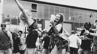 A lively outdoor gathering with people dancing and enjoying themselves. Two women in the foreground are energetically dancing and smiling, holding drinks. The background is filled with other attendees, some wearing hats and sunglasses, under a partly cloudy sky.