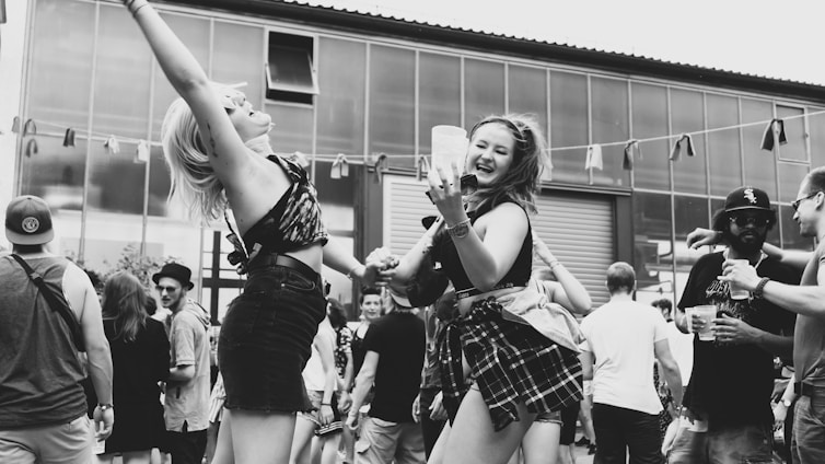 A lively outdoor gathering with people dancing and enjoying themselves. Two women in the foreground are energetically dancing and smiling, holding drinks. The background is filled with other attendees, some wearing hats and sunglasses, under a partly cloudy sky.