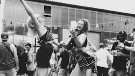 A lively outdoor gathering with people dancing and enjoying themselves. Two women in the foreground are energetically dancing and smiling, holding drinks. The background is filled with other attendees, some wearing hats and sunglasses, under a partly cloudy sky.