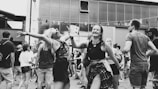 A lively outdoor gathering with people socializing and dancing. The scene includes a mix of individuals, some with drinks in hand. The setting appears to be urban, possibly at a festival or street party, with a modern building in the background.