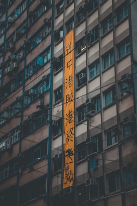 A tall building with multiple floors and identical windows, many of which feature air conditioning units. A large vertical orange banner with text hangs prominently from the building.