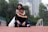 A couple is sitting on a running track with one person sitting cross-legged and another hugging from behind. The background features blurred trees and tall buildings.