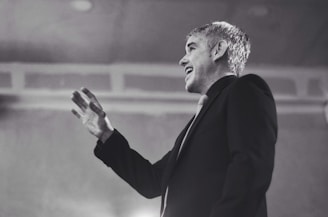 An individual is wearing a suit and gesturing with their hand as if speaking or presenting. The setting appears to be formal or professional, with a focus on the individual's expression, which conveys engagement.