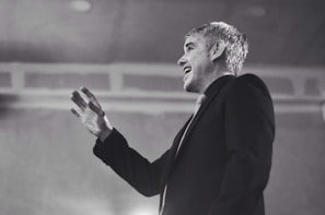 An individual is wearing a suit and gesturing with their hand as if speaking or presenting. The setting appears to be formal or professional, with a focus on the individual's expression, which conveys engagement.