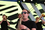 A group of musicians performing on stage, with one playing the saxophone in the foreground. They are wearing sunglasses and black shirts. In the background, other band members are playing brass instruments like trumpets and trombones. The stage backdrop features a geometric pattern with bright lighting.