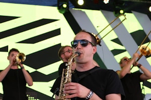 A group of musicians performing on stage, with one playing the saxophone in the foreground. They are wearing sunglasses and black shirts. In the background, other band members are playing brass instruments like trumpets and trombones. The stage backdrop features a geometric pattern with bright lighting.
