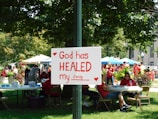 A group of people is gathered in a park-like setting with several tents and tables. In the foreground, a sign attached to a post reads 'God has HEALED my family' with red hearts decorating it. The people in the background are wearing red shirts, and the area is surrounded by green trees and plants.