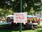 A group of people is gathered in a park-like setting with several tents and tables. In the foreground, a sign attached to a post reads 'God has HEALED my family' with red hearts decorating it. The people in the background are wearing red shirts, and the area is surrounded by green trees and plants.