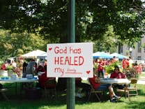 A group of people is gathered in a park-like setting with several tents and tables. In the foreground, a sign attached to a post reads 'God has HEALED my family' with red hearts decorating it. The people in the background are wearing red shirts, and the area is surrounded by green trees and plants.