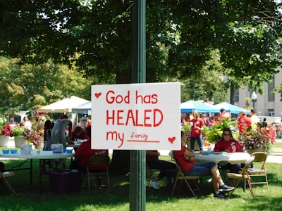 A group of people is gathered in a park-like setting with several tents and tables. In the foreground, a sign attached to a post reads 'God has HEALED my family' with red hearts decorating it. The people in the background are wearing red shirts, and the area is surrounded by green trees and plants.