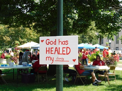 A group of people is gathered in a park-like setting with several tents and tables. In the foreground, a sign attached to a post reads 'God has HEALED my family' with red hearts decorating it. The people in the background are wearing red shirts, and the area is surrounded by green trees and plants.