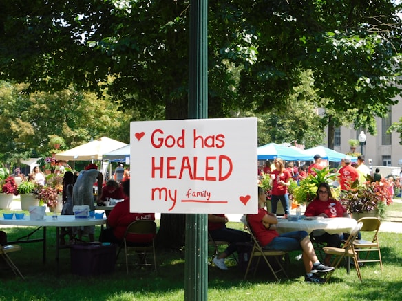 A group of people is gathered in a park-like setting with several tents and tables. In the foreground, a sign attached to a post reads 'God has HEALED my family' with red hearts decorating it. The people in the background are wearing red shirts, and the area is surrounded by green trees and plants.
