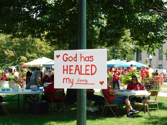 A group of people is gathered in a park-like setting with several tents and tables. In the foreground, a sign attached to a post reads 'God has HEALED my family' with red hearts decorating it. The people in the background are wearing red shirts, and the area is surrounded by green trees and plants.
