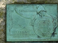 A metal plaque embedded in stone with an artistic depiction of a figure in classical armor holding a shield. The shield bears an inscription of 'Patriotism and Devotion' with stars and stripes. Above the figure's arm, 'In Memoriam' is inscribed. The text below describes the USS Maine, destroyed in Havana Harbor on February 15th, 1898, and notes the plaque is cast from the metal recovered from the ship.