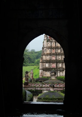 A historical temple with intricate carvings is viewed through an archway. The temple is surrounded by lush greenery, and the architecture displays detailed sculptures and patterns in stone.