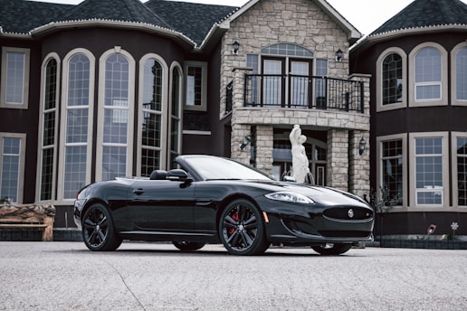 A sleek black convertible sports car is parked in front of a luxurious mansion. The mansion features large, arched windows and stone facade, enhancing the grandeur of the setting. A white statue stands at the entrance, adding a classical touch to the scene. The car's shiny exterior and stylish design are highlighted against the backdrop of the upscale building.